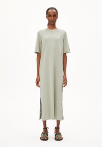 Hellgrünes, knielanges Kleid mit kurzen Ärmeln, seitlichen Schlitzen und einem minimalistischen Design. Kombiniert mit schwarzen Sandalen mit farbigen Akzenten.