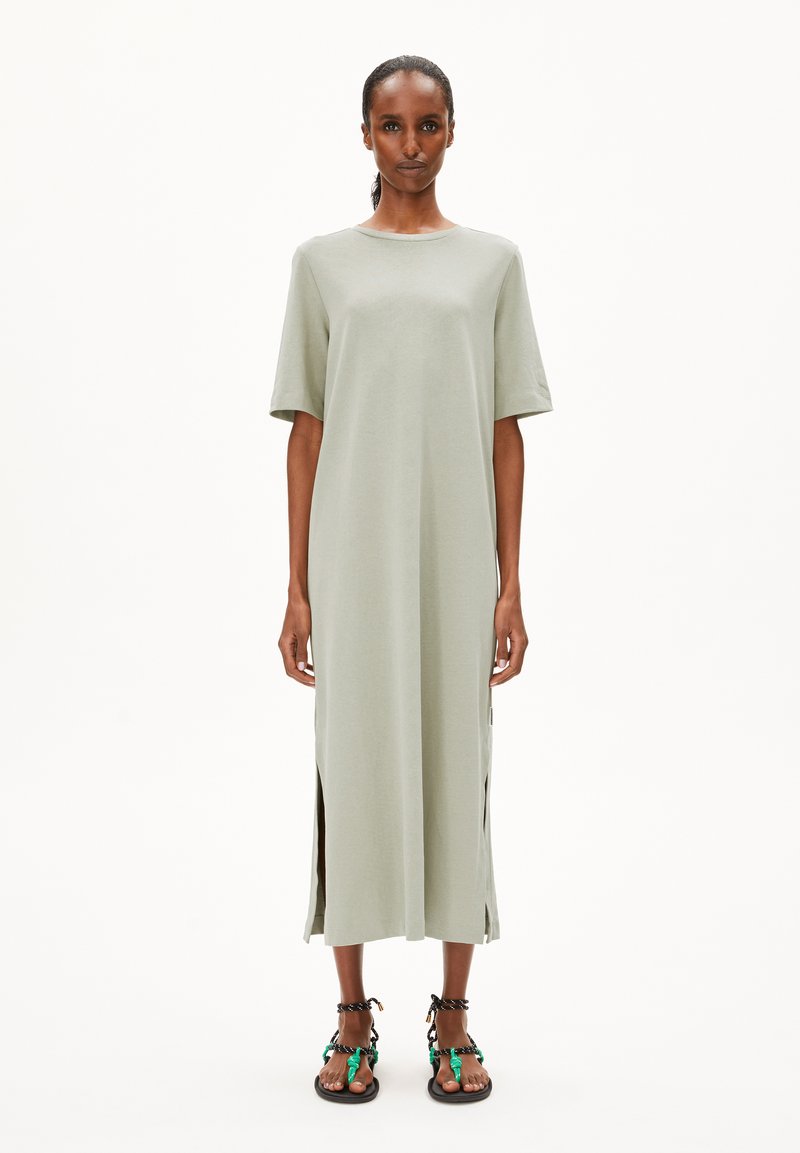 Hellgrünes, knielanges Kleid mit kurzen Ärmeln, seitlichen Schlitzen und einem minimalistischen Design. Kombiniert mit schwarzen Sandalen mit farbigen Akzenten.