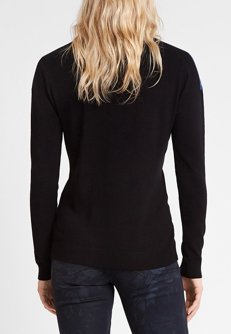 Osoba z długimi blond włosami, ubrana w dopasowany czarny top z długim rękawem i ciemne spodnie w wzory, pokazana od tyłu.