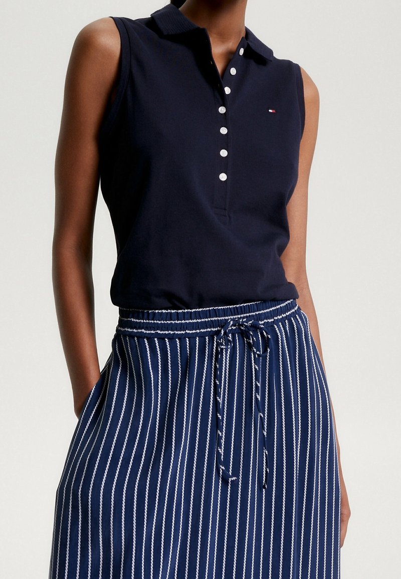 Polo sans manches bleu marine avec des boutons blancs, associé à une jupe rayée verticale bleu marine et blanche avec cordon.