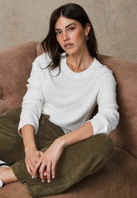 Witte gebreide trui met geribbelde manchetten, gecombineerd met olijfgroene corduroy broek. Model zit casual op een gestructureerde beige bank.