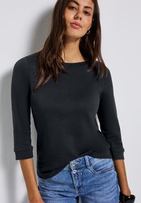 Schwarzes Langarmoberteil mit Rundhalsausschnitt. Aus glattem Stoff gefertigt, mit einem lässigen Schnitt und subtilen Saumdetails. Kombiniert mit hellblauen Jeans.