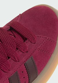 Gros plan sur une basket en daim rouge avec des lacets épais assortis, des bandes en cuir brun en zigzag et une semelle en caoutchouc marron clair.