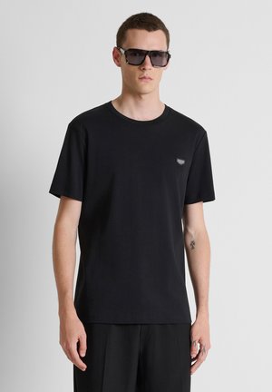 Jeune homme portant un t-shirt noir, un short noir, des lunettes de soleil carrées surdimensionnées, avec des cheveux courts et un petit tatouage sur l'avant-bras, debout devant un fond uni.