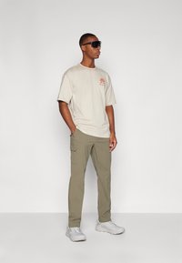 Camiseta de algodón beige con cuello redondo y logo naranja, combinada con pantalones cargo oliva y zapatillas gris claro. Llevada con gafas de sol negras.