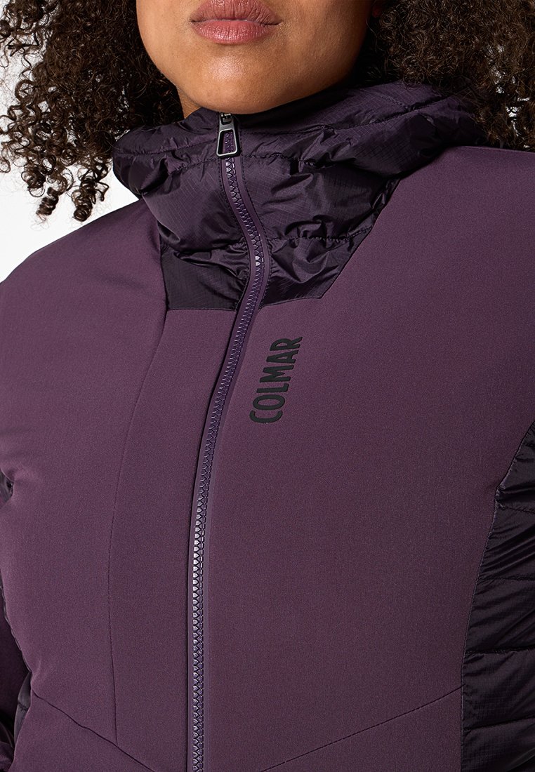 Giacca a vento viola con colletto alto, chiusura con zip e logo del marchio. Presenta un tessuto liscio e lucido con texture e sezioni a contrasto.