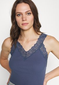 Marineblå camisole med ribbestruktur, med en delikat blonderkant langs V-halsen og stroppene, som viser intrikate blomsterdesign.
