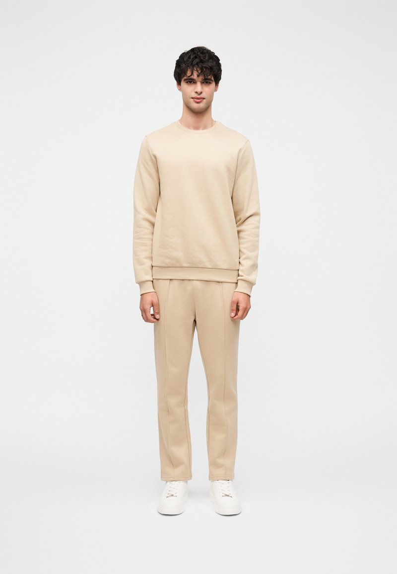 Μπεζ φούτερ και αντίστοιχο παντελόνι φόρμας από απαλό ύφασμα. Διαθέτουν στρογγυλή λαιμόκοψη και ελαστικά μανίκια. Φοριούνται με λευκά αθλητικά παπούτσια.