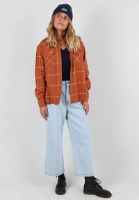 Chemise en flanelle à carreaux orange avec deux poches poitrine et des coutures blanches, portée par-dessus un haut navy, assortie à un jean large bleu clair et des bottes noires.