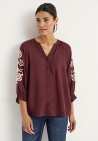 Bordignonin nappipaita, jossa on V-pääntie, kolme neljäsosaa hihat ja valkoista kukka-embroiderya hihoissa. Pehmeä, sileä kangas, jossa on kerättyjä hihansuita.