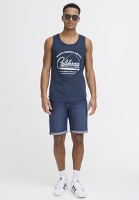 Mornarska majica brez rokavov z belim grafičnim besedilom, kavbojke s prevrnjenimi manšetami, bele nogavice in črno-bele športne čevlje, dodatno opremljene z sončnimi očali.