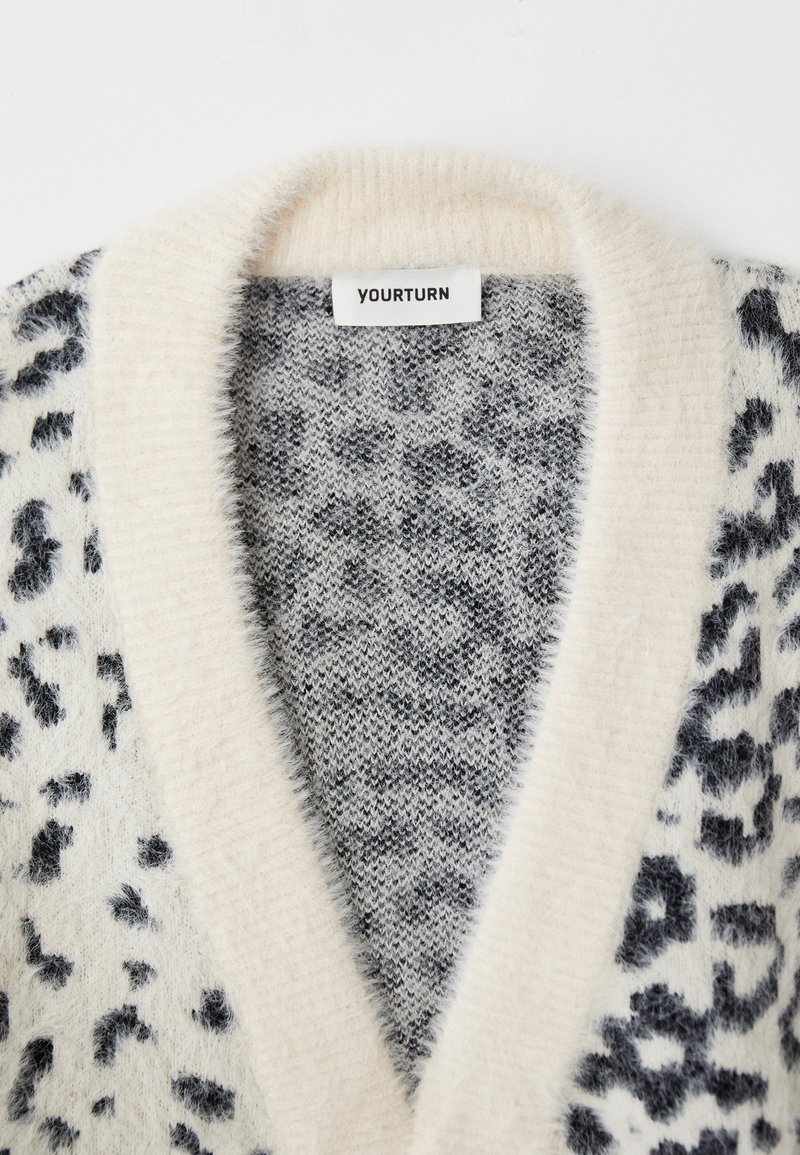 Mjukt och fluffigt cardigan med svart och grå leopardmönster samt krämfärgad sjalkrage, visar "YOURTURN" märklapp.