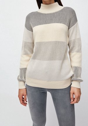 Femme portant un pull à col roulé en maille rayée crème et noire et un pantalon gris, debout devant un fond uni.