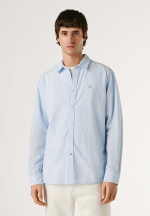 Junger Mann trägt ein hellblaues, langärmliges Hemd mit dünnen vertikalen Streifen und weiße Hosen vor einfachem Hintergrund.