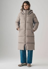 Beige gepolsterter langer Mantel mit Kapuze, mit Reißverschluss, seitlichen Taschen und horizontalem Steppmuster, kombiniert mit weiten Jeans.