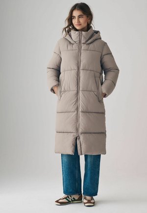 Beige vadderad långjacka med huva, som har dragkedja, sidfickor och horisontellt kviltat mönster, tillsammans med vida jeans.