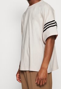 Ljusbeige bomull t-shirt med korta ärmar, med svarta trippla ränder på vänster arm och en avslappnad passform. Sidoklyftor vid fållen.