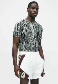 Grafiska īsroku krekls melnā un baltā viļņu rakstā, kombinēts ar baltām šortām ar bordo un zaļiem akcentiem un logotipu.