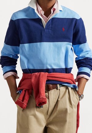 Mand iført blåstribet rugbyshirt med lyserød knaplukket krave synlig, rød sweatshirt bundet om livet, beige bukser og et armbåndsur.