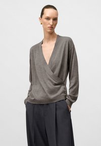 Donna che indossa un maglione grigio a maniche lunghe a wrap con bottoni sul lato e pantaloni grigi scuri plissettati e larghi, con le mani nelle tasche.