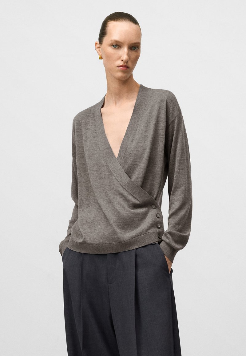 Donna che indossa un maglione grigio a maniche lunghe a wrap con bottoni sul lato e pantaloni grigi scuri plissettati e larghi, con le mani nelle tasche.
