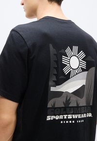 Μαύρο βαμβακερό μπλουζάκι με γραφικό τύπωμα στην πλάτη, που απεικονίζει τον ήλιο και τοπίο σε αποχρώσεις του γκρι, με το κείμενο «Columbia Sportswear Co.».