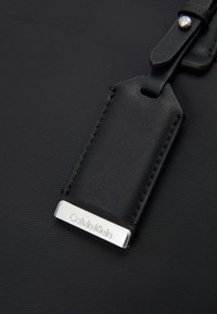 Étiquette de bagage en cuir noir avec plaque métallique argentée gravée "Calvin Klein" attachée à une surface en tissu noir.