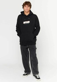 Sweat-shirt à capuche noire avec une poche avant et un texte graphique coloré. Associé à un jean noir ample et des baskets noires.