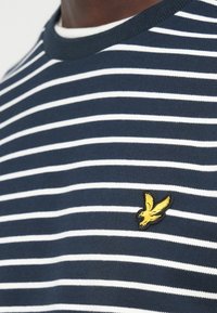 Μπλε ναυτικό και άσπρο ριγέ μπλουζάκι με στρογγυλή λαιμόκοψη και μικρό κεντητό κίτρινο λογότυπο πουλιού στην αριστερή πλευρά του στήθους.