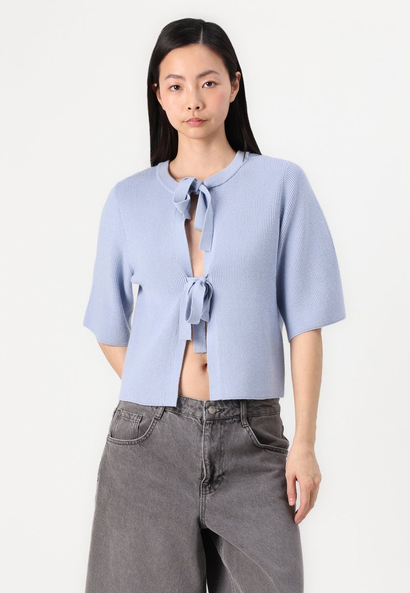 Jasnoniebieski, dzianinowy, krótki kardigan z krótkimi rękawami, otwarty z przodu i z zapięciem na sznurek. Połączony z luźnymi szarymi dżinsami.