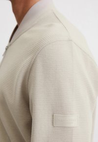 Beige textured sweater with a ribbed pattern, featuring a zip and a branded label on the left shoulder; made from a soft knit material.