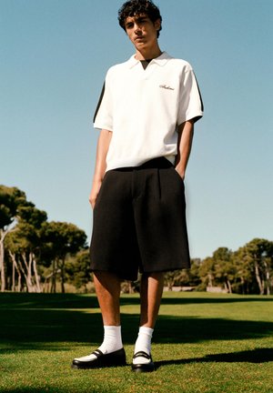 Egy fiatal férfi áll a fűben, fehér, fekete csíkos galléros inget, fekete bő rövidnadrágot, fehér zoknit és fekete mokaszint viselve tiszta ég alatt.