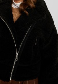 Musta tekoturkishenkinen takki vetoketjulla, suurella kauluksella ja sivutaskulla. Ominaisuudet: pehmeä tekstuuri ja rento istuvuus.