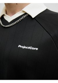 Sort polo-shirt med vertikale striber, hvid krave og sølvfarvet kædedetalje. Har "ProjectCore" logo i hvidt på brystet.