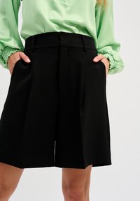 Czarne spodenki o dopasowanym kroju z prostymi nogawkami, detalem w postaci fałdy w talii i kieszeniami po bokach; w zestawie z jasnoniebieską bluzką z falbankami przy mankietach.