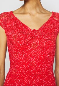 Femme portant une robe rouge à pois blancs avec des détails en dentelle, avec un nœud sur la poitrine et des manches courtes.