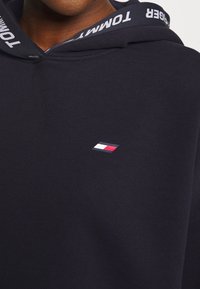 Sudadera con capucha de color azul marino oscuro, hecha de algodón. Presenta un logo en el pecho en rojo, blanco y azul. La capucha tiene una banda interior de logo en contraste.