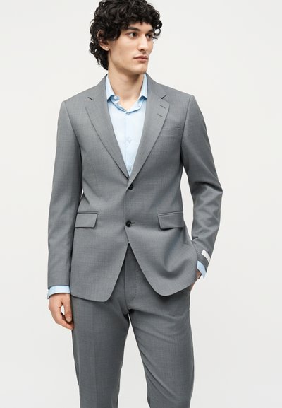 Νέος άνδρας με σγουρά σκουρόχρωμα μαλλιά, ντυμένος με γκρι κοστούμι και ανοιχτό γαλάζιο πουκάμισο, στέκεται με ένα χέρι στην τσέπη, κοιτώντας δεξιά.