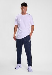 Witte T-shirt met korte mouwen en zwart logo, gecombineerd met marineblauwe trainingsbroek met een gestreepte detail aan de zijkant en witte sneakers.