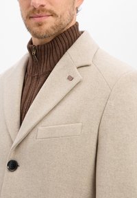 Man in een beige wollen jas met zwarte knopen en een Britse vlagspeld op een bruine geribde turtleneck sweater.