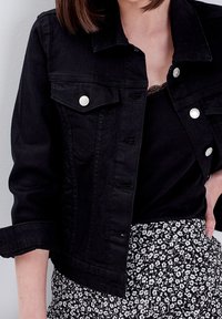 Zwarte denimjack met zilveren knopen, twee voorkezen, en opgerolde mouwen, gecombineerd met een zwart top en een rok met bloemmotief.