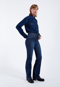 Camicia in denim blu scuro con due tasche sul petto, abbinata a jeans a zampa d'elefante blu scuro a vita alta. Il modello indossa scarpe con suola platform nera.