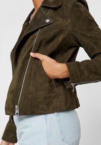 Jasnozielona zamszowa kurtka motocyklowa z srebrnymi detalami, zapinanymi na zamek kieszeniami i dopasowanym krojem. Posiada szeroki kołnierz oraz fakturowaną powierzchnię.