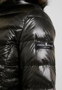 Manche de veste matelassée noire brillante avec capuche bordée de fourrure et étiquette en tissu "Frieda & Freddies New York" sur le haut du bras.