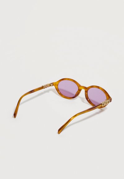 Lunettes de soleil rondes à monture écaille d'ambre avec des verres violets et des accents de logo doré sur les branches, sur un fond blanc uni.