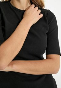Černé žebrované tričko s krátkými rukávy a kulatým výstřihem. Tkanina se zdá být měkká s přiléhavým siluetou. Předloktí spočívá na hrudníku.
