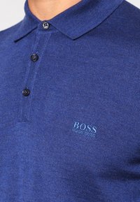 Polo bleu en tissu côtelé, avec un col à boutons, trois boutons noirs et un logo bleu clair brodé sur la poitrine.