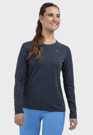 Lächelnde Frau mit langen braunen Haaren im Pferdeschwanz, die ein marineblaues langärmeliges Shirt und leuchtend blaue Hosen trägt und leicht nach rechts schaut.