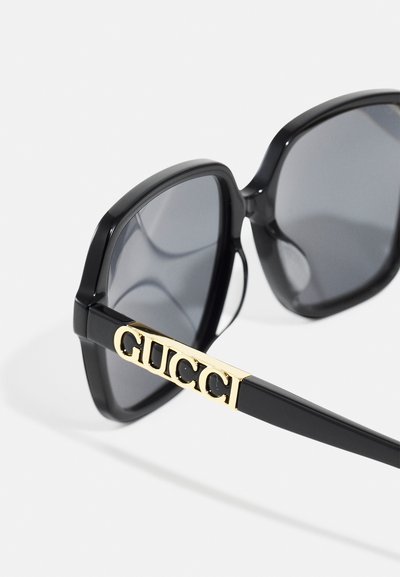 Lunettes de soleil noires rectangulaires avec des verres gris. La branche présente un accent en or avec le logo "GUCCI". Les montures sont brillantes et au design moderne.