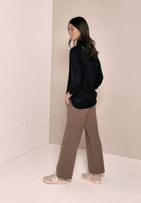 Femme en chemise noire à manches longues, pantalon large marron et baskets beige, debout de profil contre un mur beige uni.